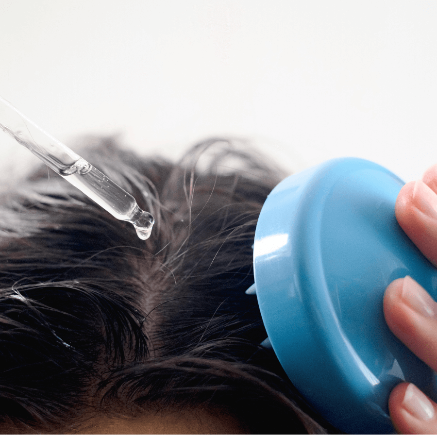woman apply oil to her scalp with a dropper in one hand and a scalp. massage brush in the other