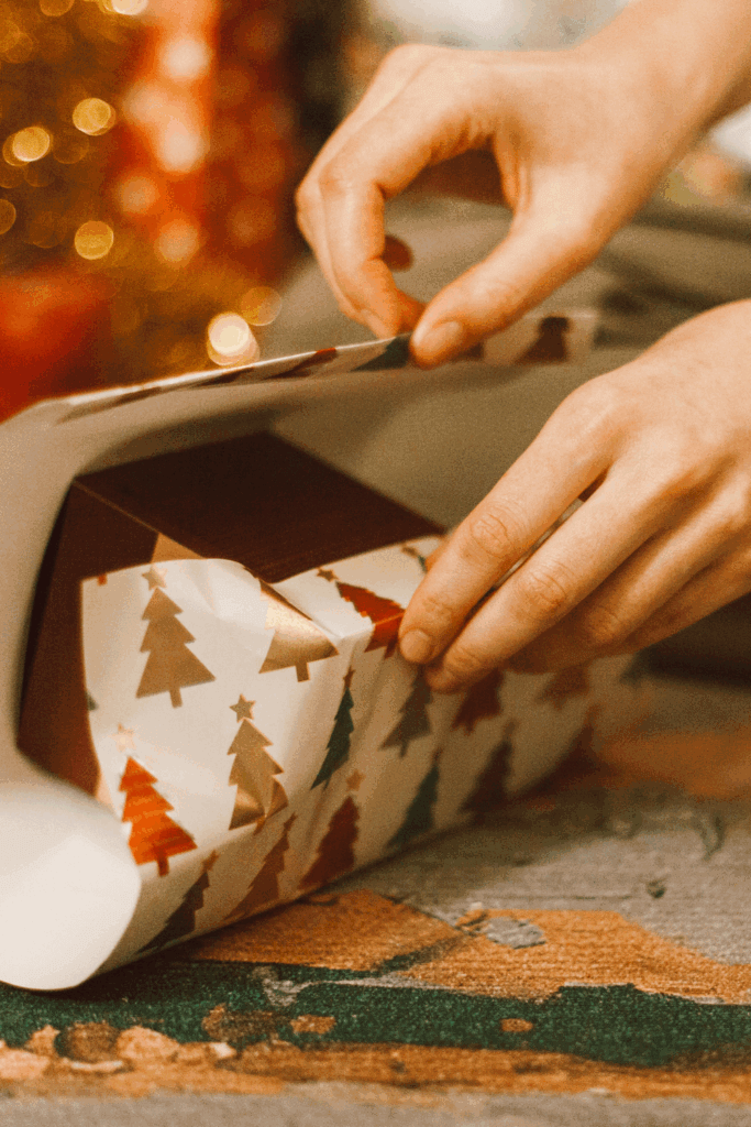 Close-up of hands carefully wrapping a gift in Christmas tree patterned wrapping paper, with a festive golden bokeh background.