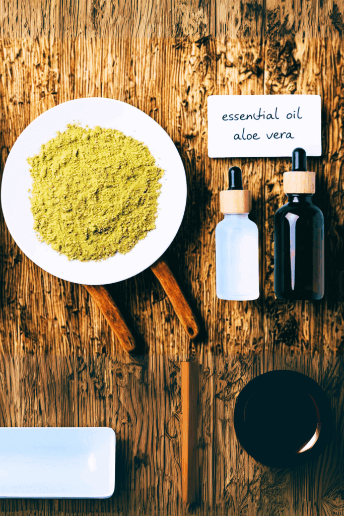Top-down view of a white plate with green herbal powder, dropper bottles labeled essential oil and aloe vera, and natural containers arranged on a rustic wooden surface.