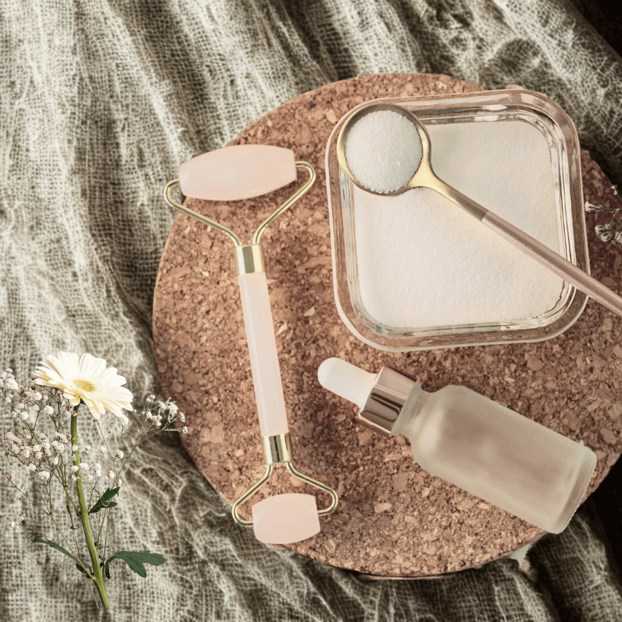 soft and calming flat lay featuring a rose quartz face roller, a glass dropper bottle with serum, and a bowl of fine powder with a gold measuring spoon. The items sit on a cork surface with a textured fabric and a single white flower nearby, creating a peaceful beauty and self care setting.