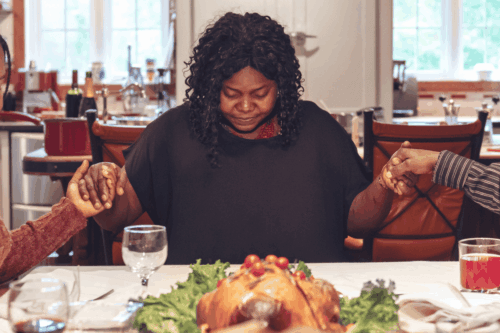 black woman bowing her head praying over the thanksgiving table while holding hands with a person on each side of her